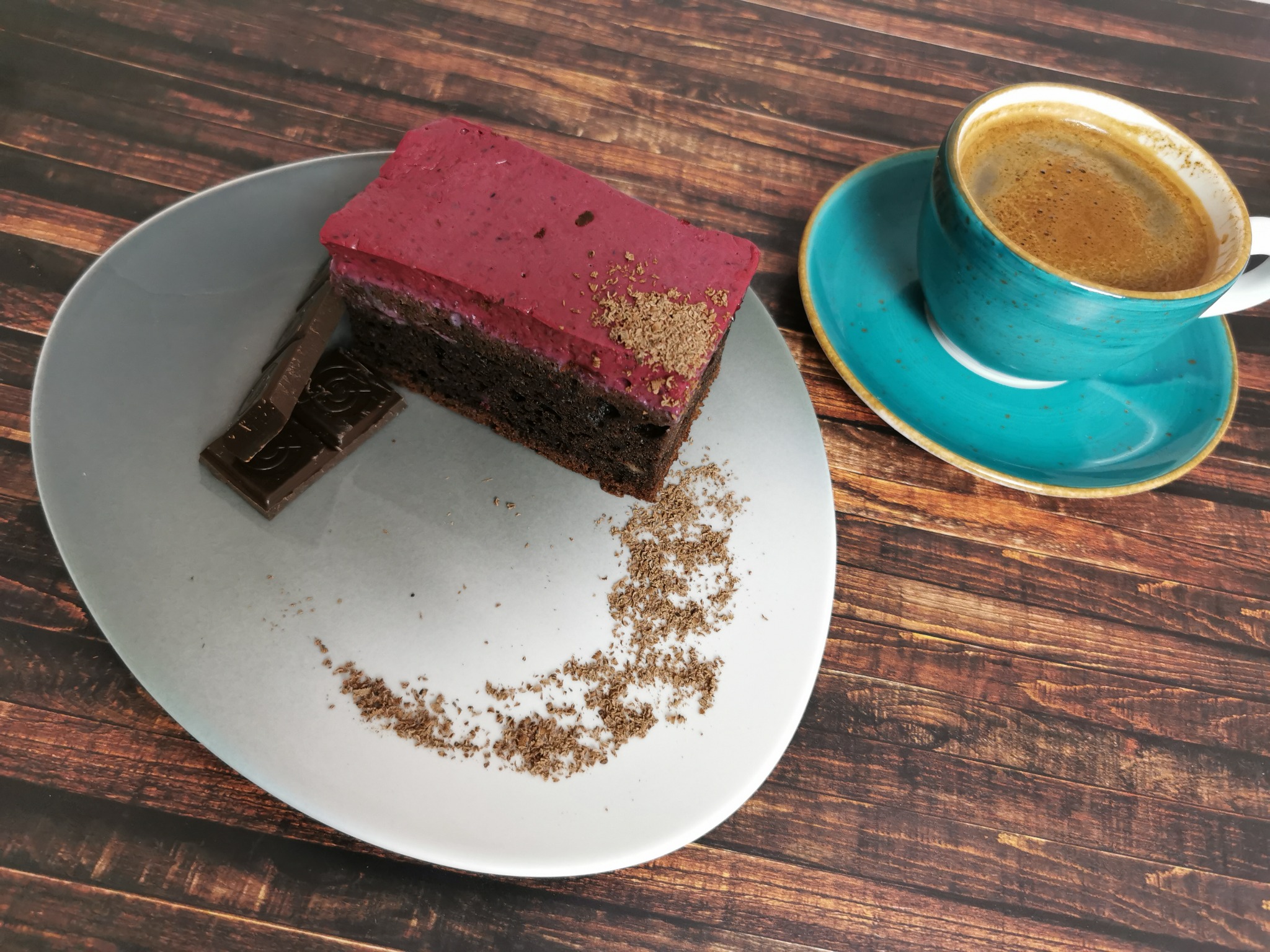 Kawałek czekoladowego ciasta z różową polewą i kawałkiem czekolady na szarym talerzu, obok turkusowa filiżanka kawy na drewnianym stole.