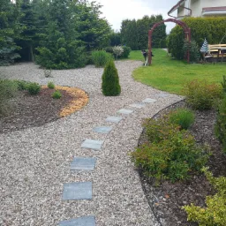 Ścieżka z płyt chodnikowych ułożona w żwirze w ogrodzie z różnorodną roślinnością, w tle widoczna pergola i wypoczynek ogrodowy.