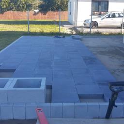 Pan Kostka - Fragment układanej nawierzchni z szarych płyt betonowych na posesji, widoczne narzędzia brukarskie, w tle dom jednorodzinny i trawnik.