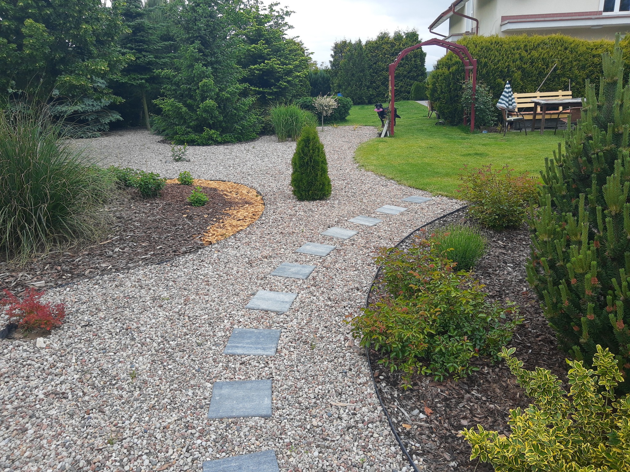 Ścieżka z płyt chodnikowych ułożona w żwirze w ogrodzie z różnorodną roślinnością, w tle widoczna pergola i wypoczynek ogrodowy.