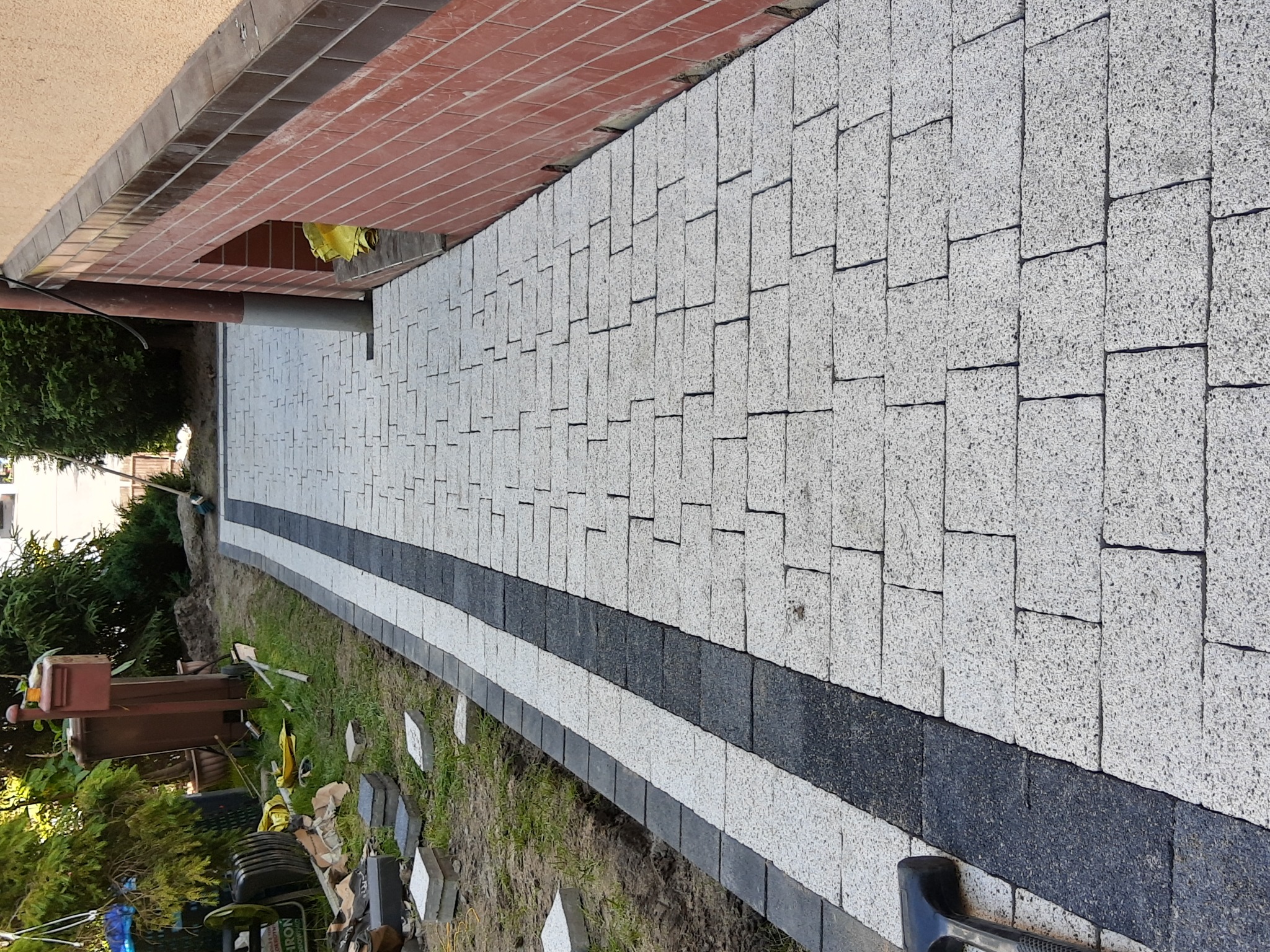 Ścieżka z jasnoszarej kostki z ciemnoszarym pasem pośrodku, widok z góry, obrzeża z ciemnej kostki, trawa po bokach, fragment budynku w tle.