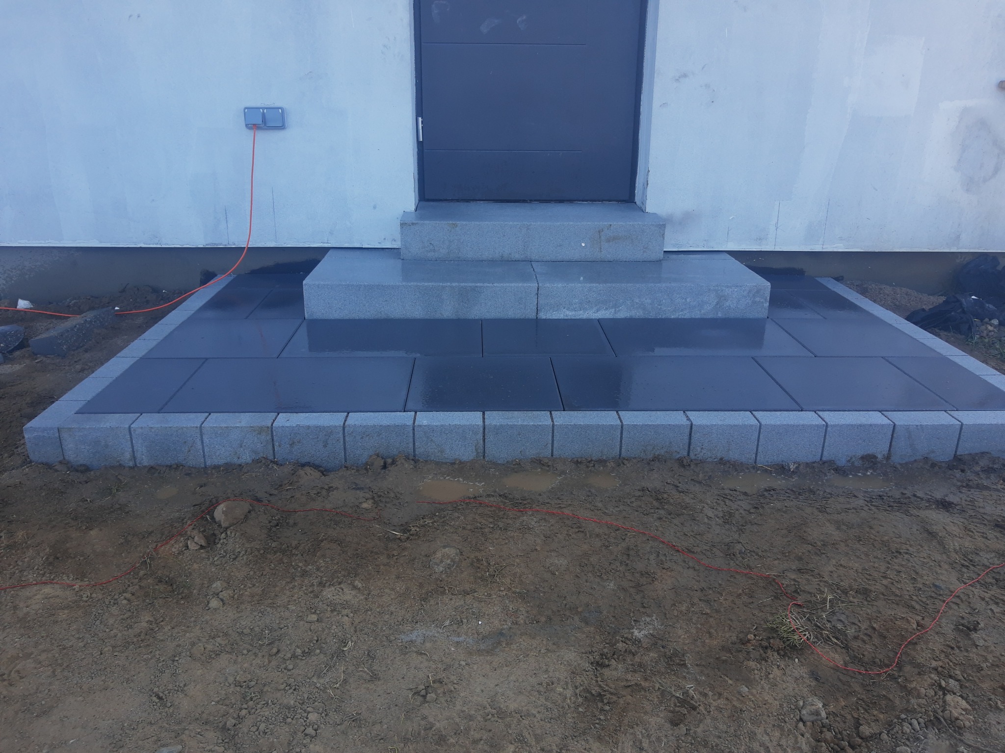 Ułożona, mokra kostka granitowa przed wejściem do budynku, otoczona brudnym terenem z widocznymi śladami po pracach ziemnych i czerwoną linką wyznaczającą obszar robót.