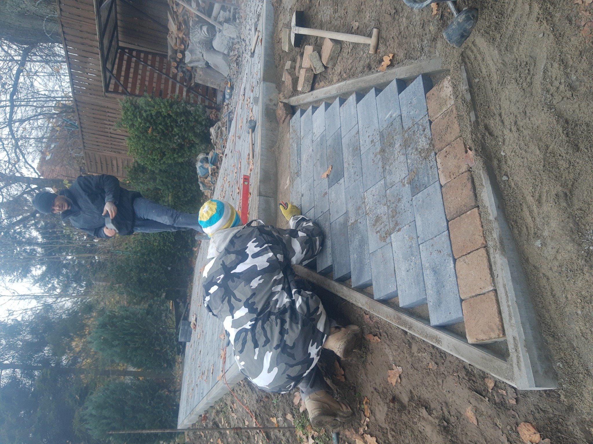 Robotnicy układają schody z kostki brukowej w ogrodzie, widoczne narzędzia i materiały budowlane, jeden z pracowników w ubraniu moro i czapce w paski.