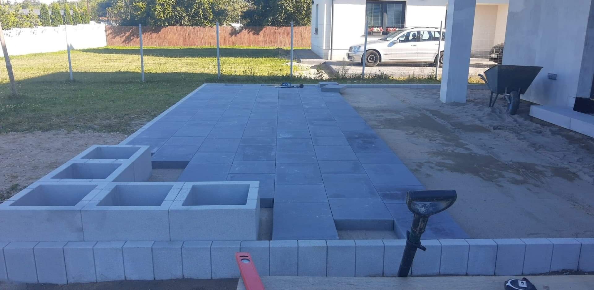 Fragment układanej nawierzchni z szarych płyt betonowych na posesji, widoczne narzędzia brukarskie, w tle dom jednorodzinny i trawnik.