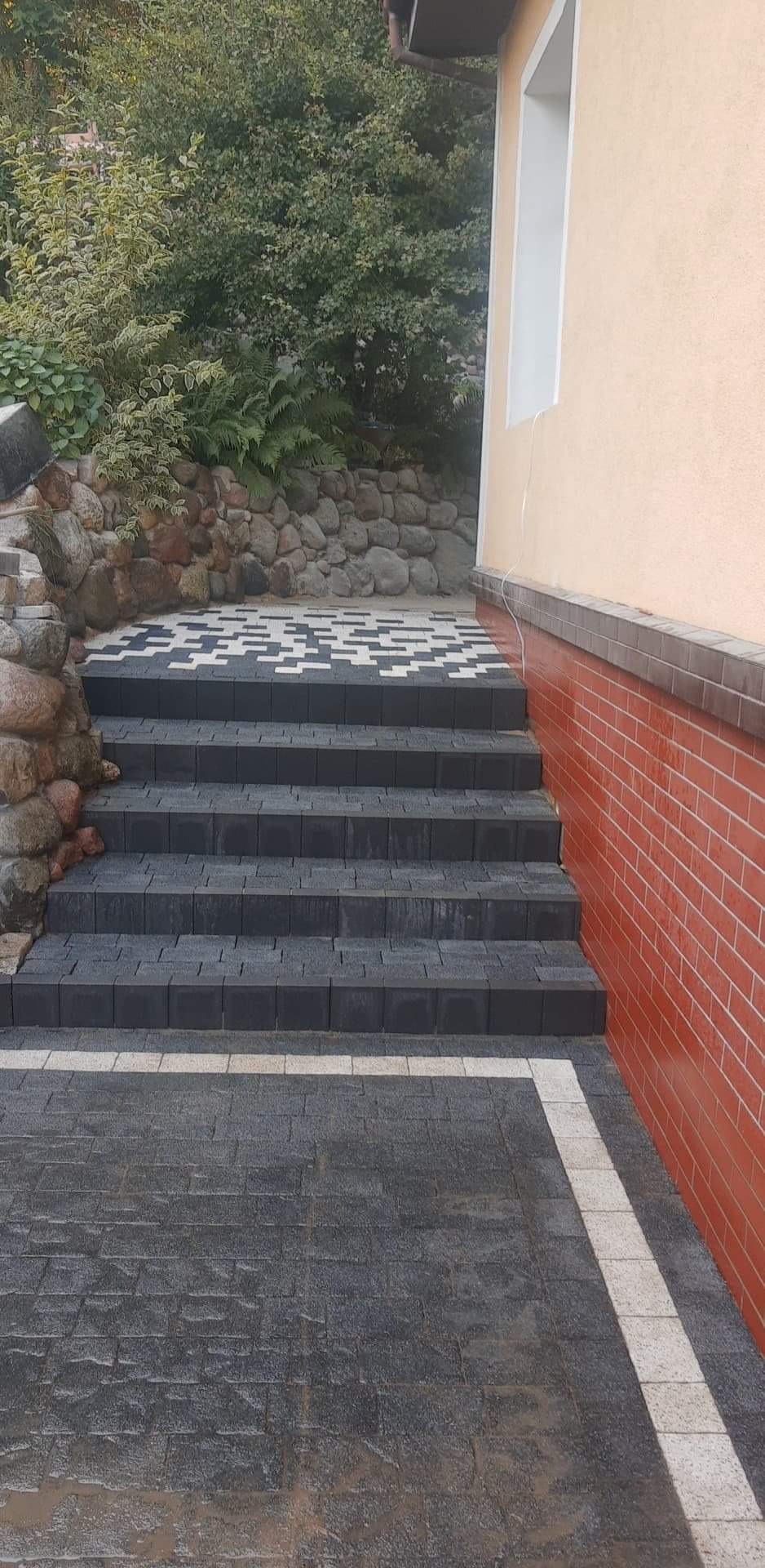 Schody zewnętrzne z ciemnej kostki brukowej z jasnymi obramowaniami, prowadzące w górę obok kamiennego murku i ściany budynku z ceglaną okładziną.