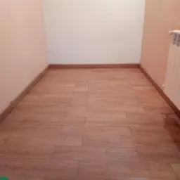 Pusta przestrzeń z panelami podłogowymi imitującymi drewno, listwy przypodłogowe z płytek ceramicznych i grzejnik żeberkowy przy ścianie.