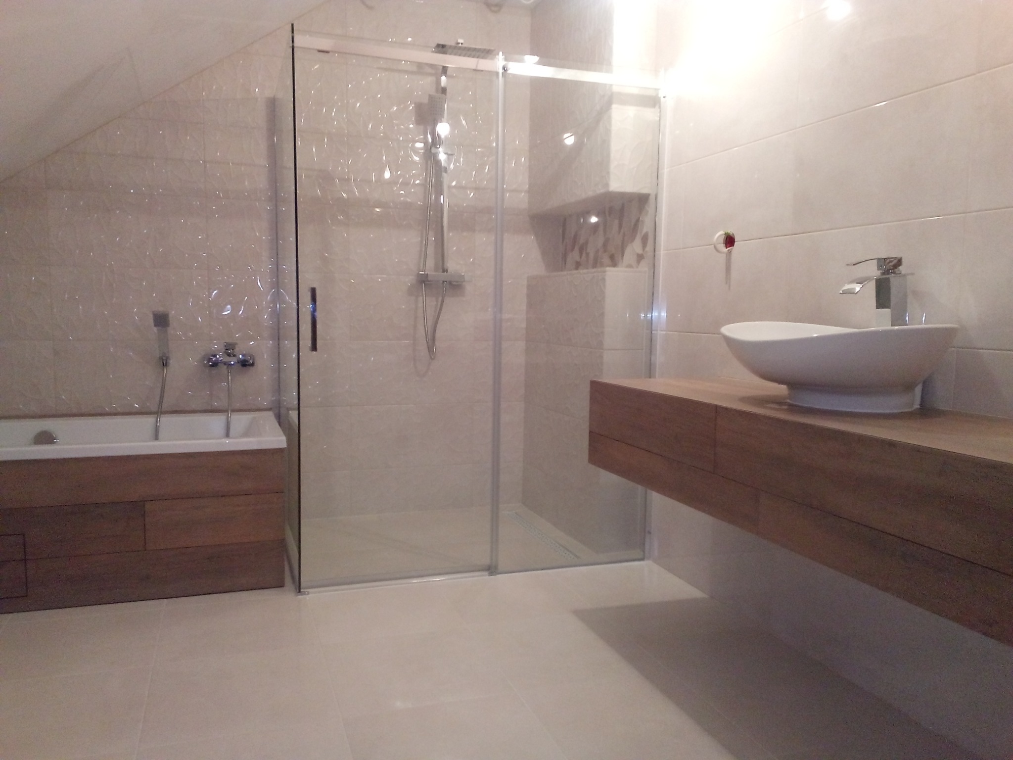 Nowoczesna łazienka z kabiną prysznicową ze szklanymi drzwiami przesuwnymi, wiszącą umywalką na drewnianym blacie i obudową wanny z drewnianymi panelami, wykończona beżowymi płytkami z delikatną...