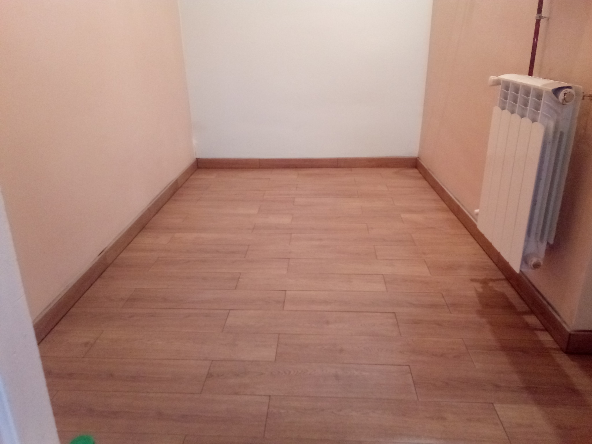 Pusta przestrzeń z panelami podłogowymi imitującymi drewno, listwy przypodłogowe z płytek ceramicznych i grzejnik żeberkowy przy ścianie.