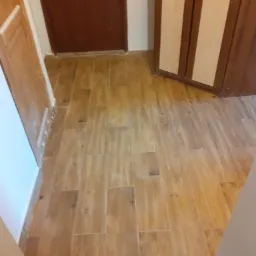 Ułożona podłoga z płytek imitujących drewno, widoczne drzwi i szafka w tle, fragment listwy przypodłogowej.