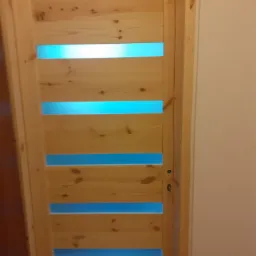 Nowoczesne drzwi wewnętrzne z surowego drewna z poziomymi, prostokątnymi prześwitami, przez które widać błękitne tło, zamontowane w ościeżnicy.