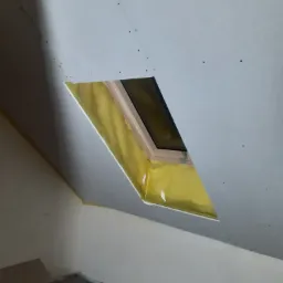 Montaż płyty karton gips wokół okna dachowego, widoczne wkręty mocujące i izolacja termiczna w otworze okiennym.