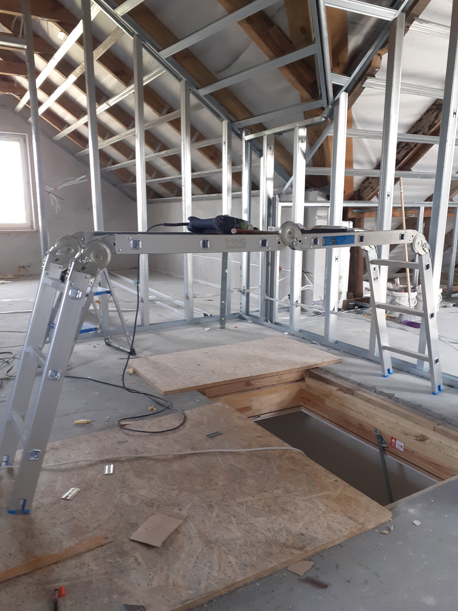 Stelaż z profili stalowych na poddaszu, widoczne drewniane elementy konstrukcji dachu, drabina platformowa z wiertarką, otwór w podłodze zabezpieczony płytą.