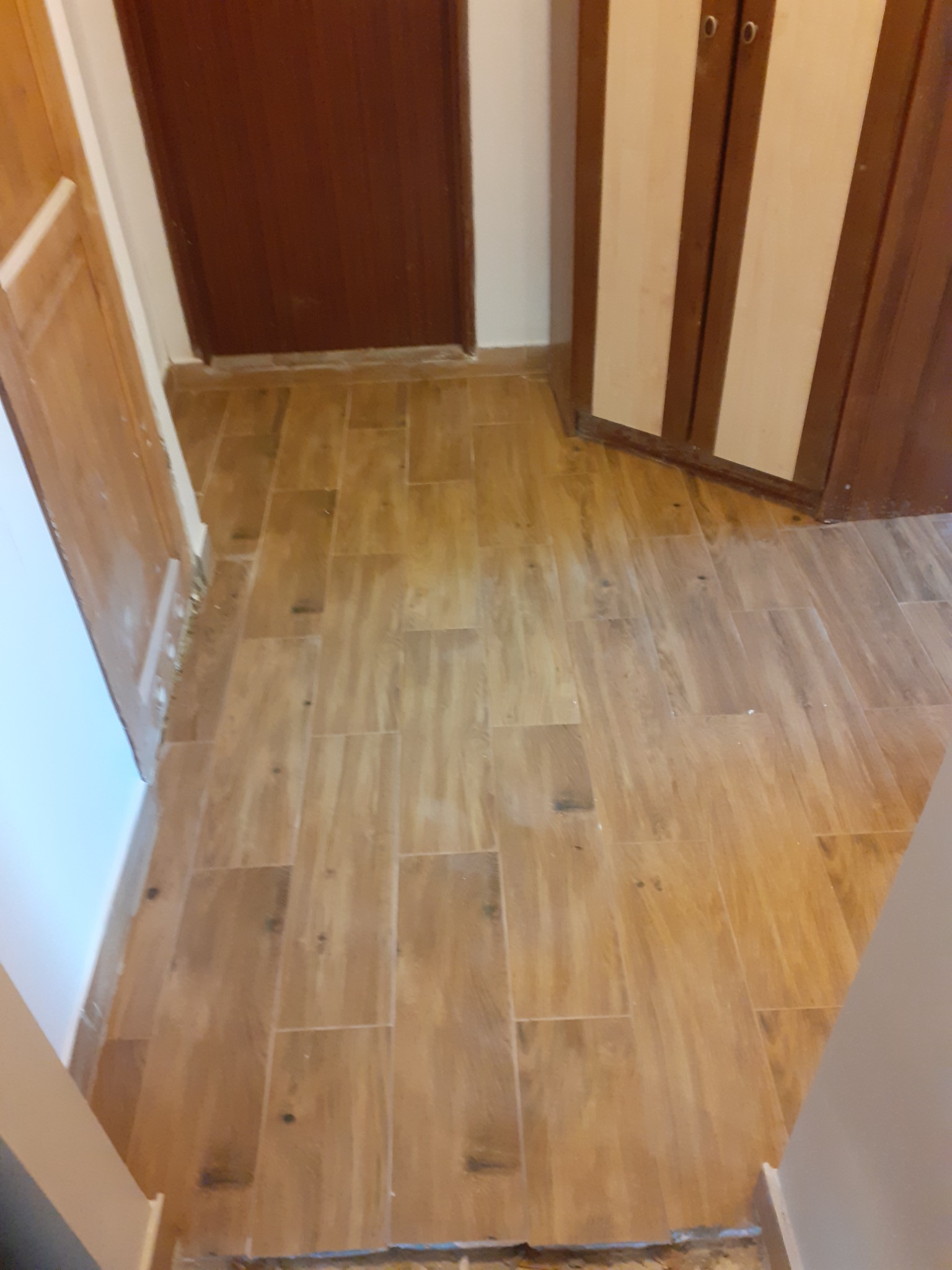 Ułożona podłoga z płytek imitujących drewno, widoczne drzwi i szafka w tle, fragment listwy przypodłogowej.