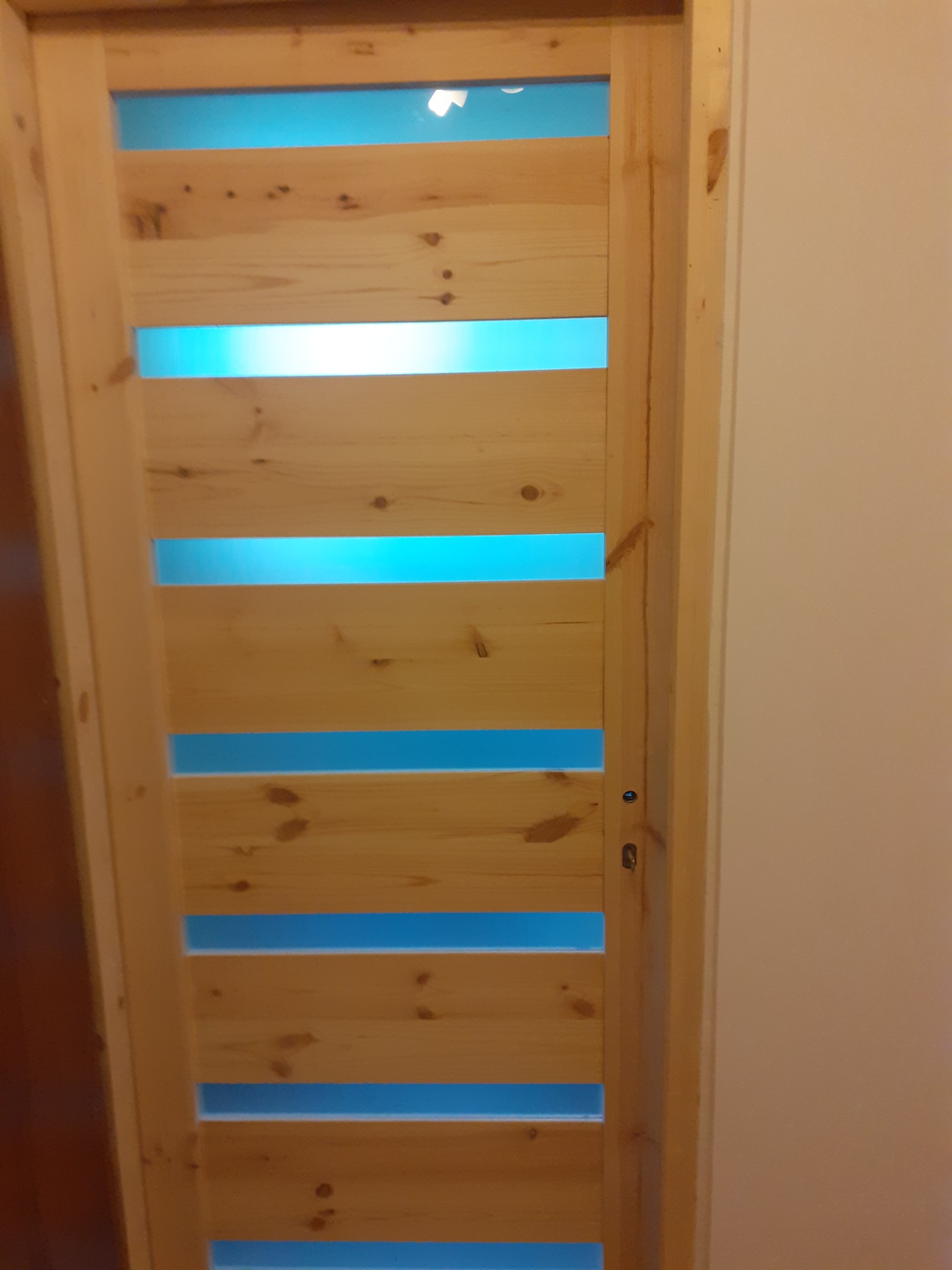 Nowoczesne drzwi wewnętrzne z surowego drewna z poziomymi, prostokątnymi prześwitami, przez które widać błękitne tło, zamontowane w ościeżnicy.