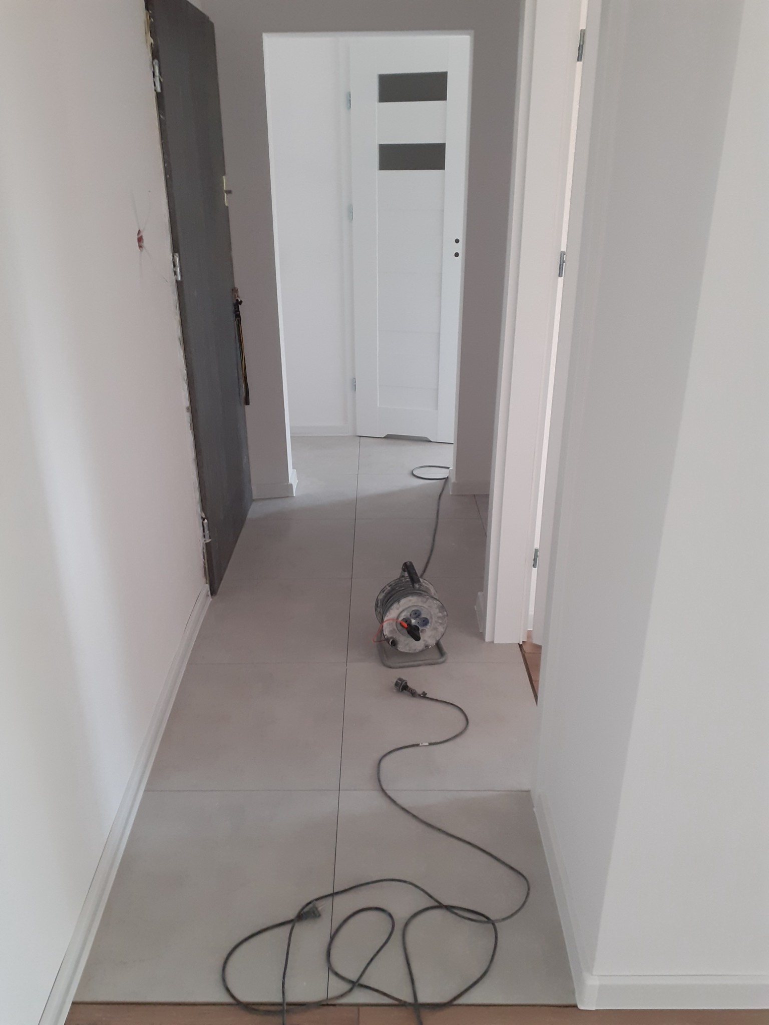 Korytarz w trakcie prac wykończeniowych z widocznym agregatem prądotwórczym na szarych płytkach podłogowych, ściany świeżo pomalowane na biało, widoczne niedoskonałości na ścianie po lewej stronie.