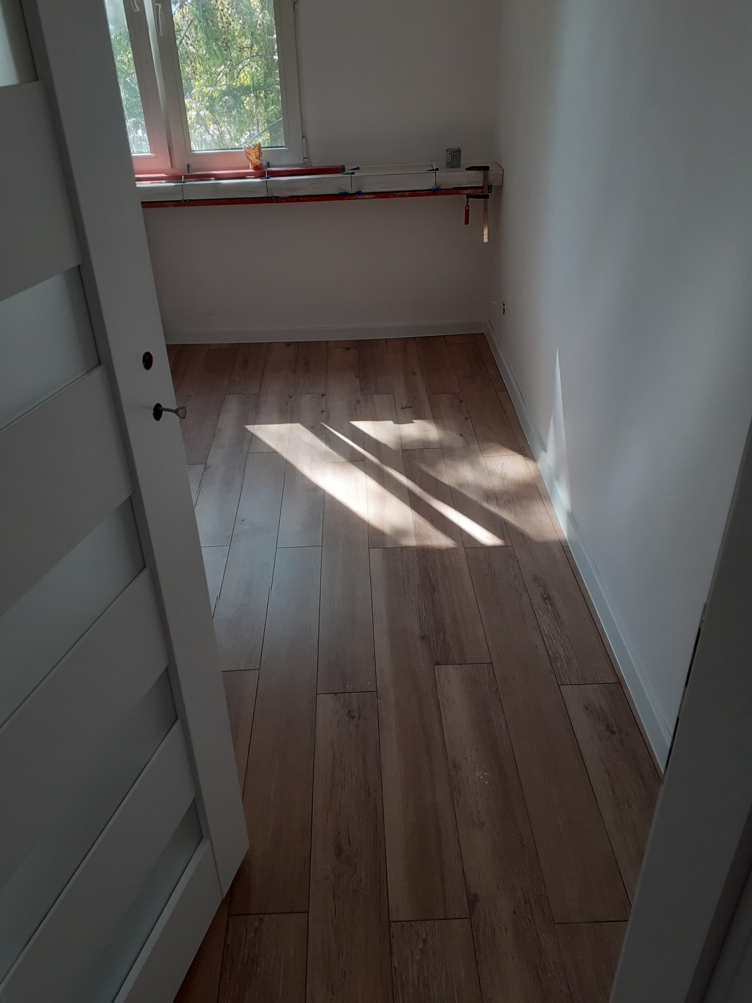 Jasne panele podłogowe ułożone w pustym, pomalowanym na biało pokoju z widocznym fragmentem okna i otwartymi, białymi drzwiami.