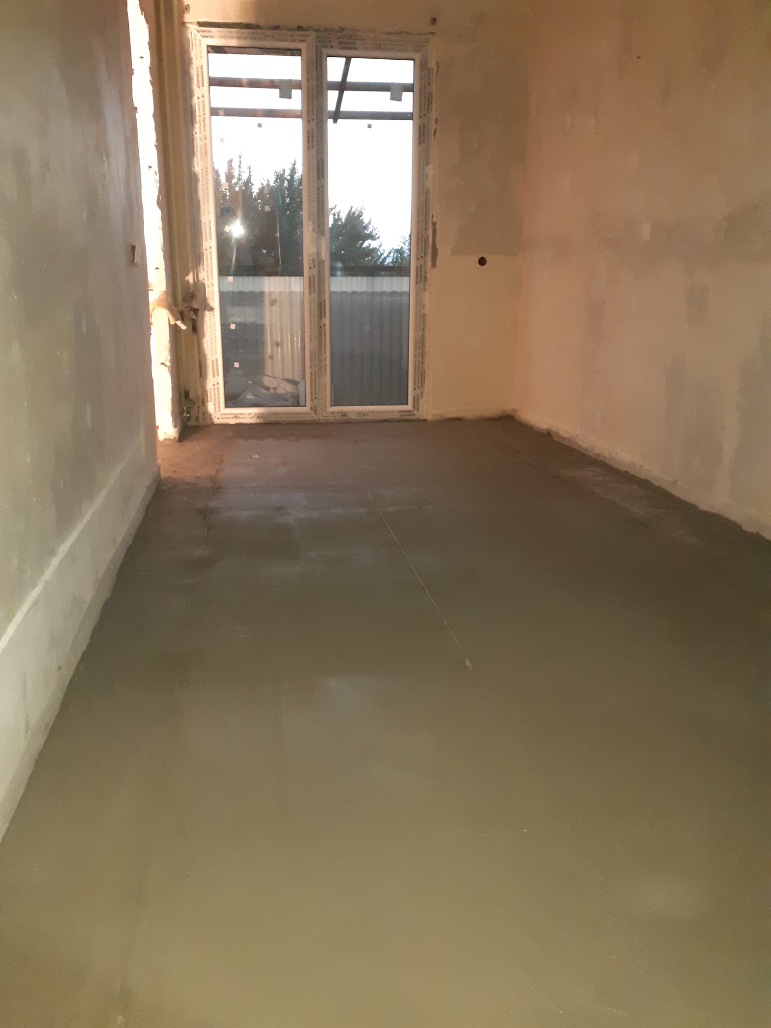 Wylewka samopoziomująca na podłodze w pomieszczeniu w trakcie schnięcia, widoczne ślady po narzędziach i sznurek wyznaczający poziom, ściany przygotowane pod malowanie, okno balkonowe z widokiem...