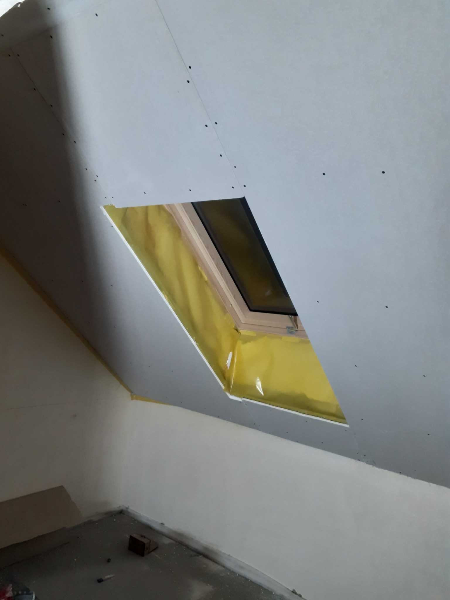 Montaż płyty karton gips wokół okna dachowego, widoczne wkręty mocujące i izolacja termiczna w otworze okiennym.