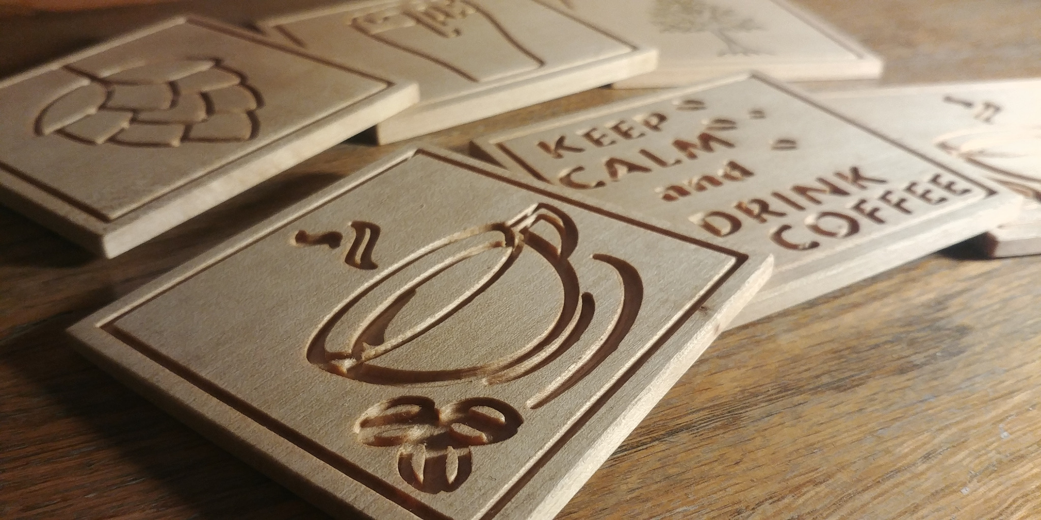 Drewniane podkładki z wygrawerowanymi wzorami: filiżanka kawy, napis 'Keep Calm and Drink Coffee', szyszka chmielu i drzewo.