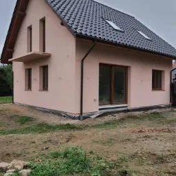 Nowo otynkowany dom jednorodzinny z ciemną dachówką, widoczne okna dachowe i mały balkon, teren wokół domu częściowo zagospodarowany.