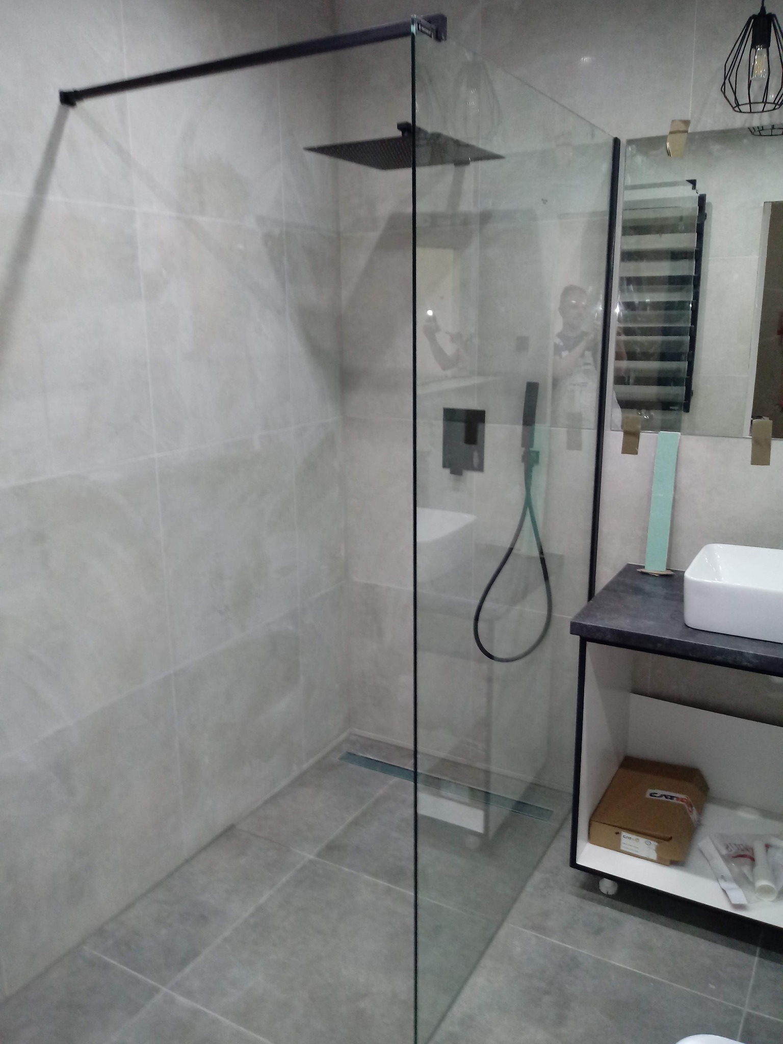 Nowoczesna łazienka z kabiną prysznicową typu walk-in, wykończona szarą płytką ceramiczną, z czarnymi detalami armatury i oświetlenia, oraz elementami wyposażenia w postaci umywalki nablatowej...