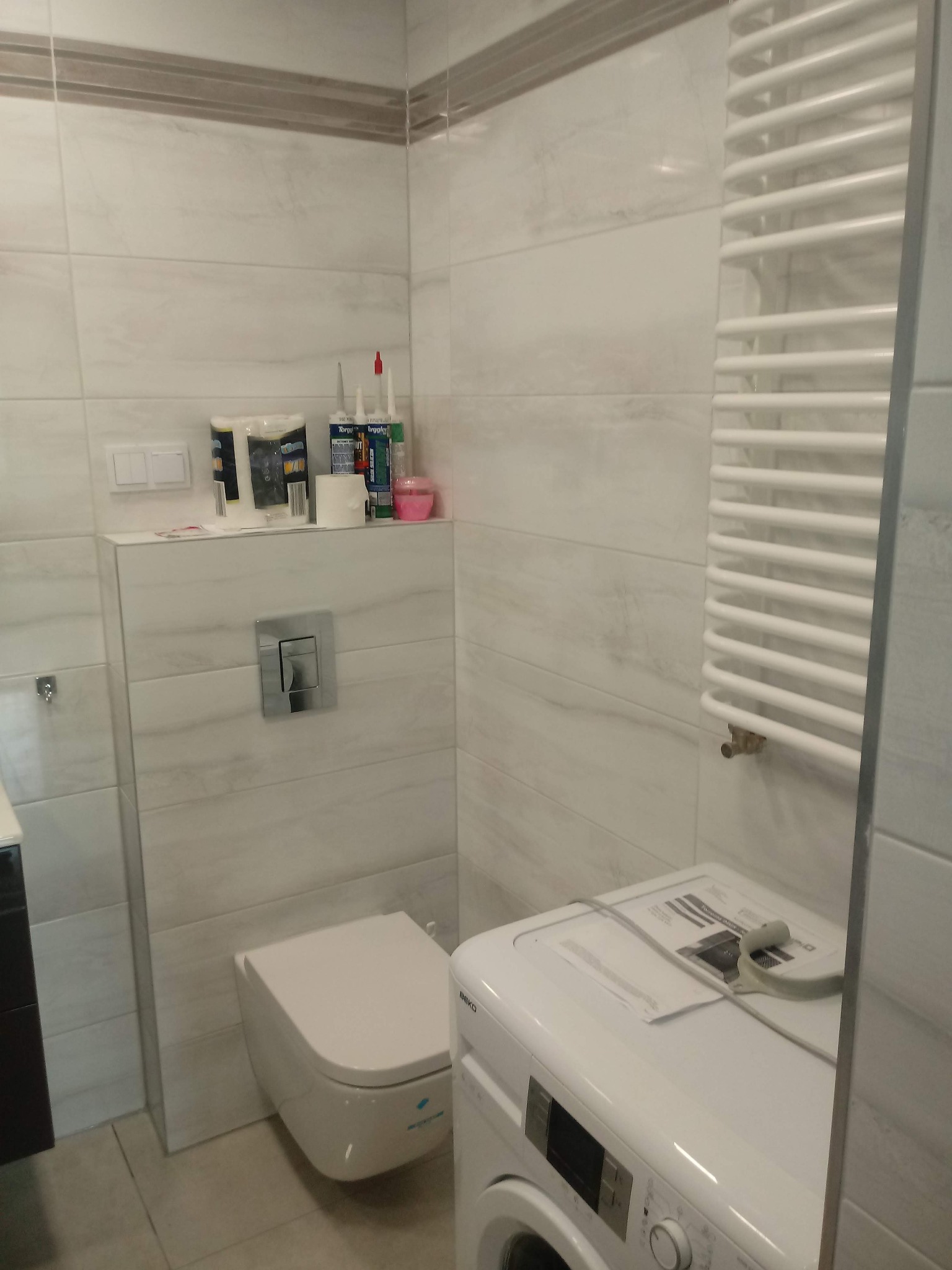 Nowoczesna łazienka z podwieszaną toaletą, grzejnikiem drabinkowym i pralką Beko, wykończona płytkami imitującymi marmur, z widocznymi materiałami budowlanymi na półce.