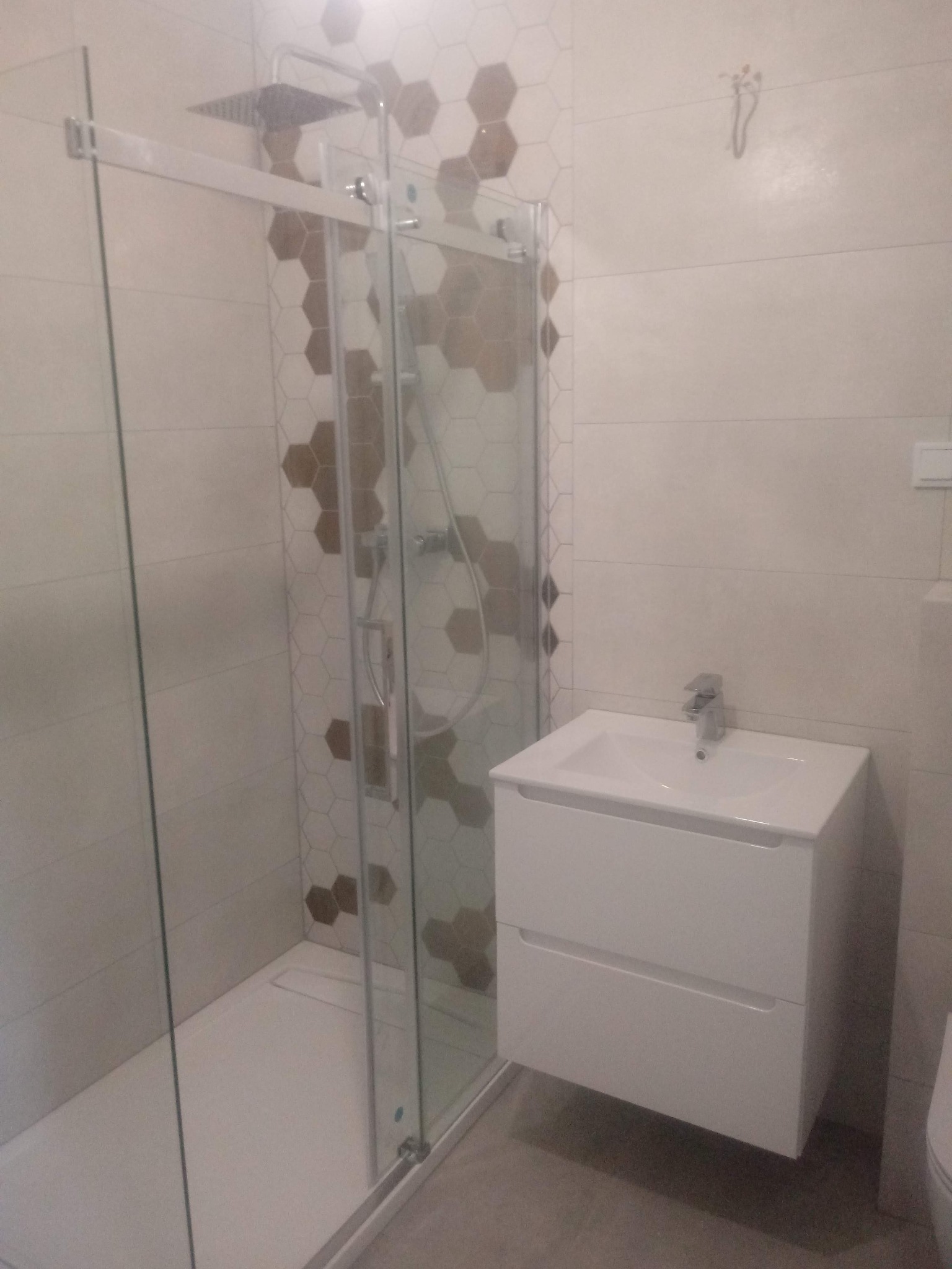 Nowoczesna łazienka z kabiną prysznicową typu walk-in z deszczownicą, umywalką z szafką w kolorze białym i heksagonalnymi płytkami dekoracyjnymi w odcieniach beżu i brązu.