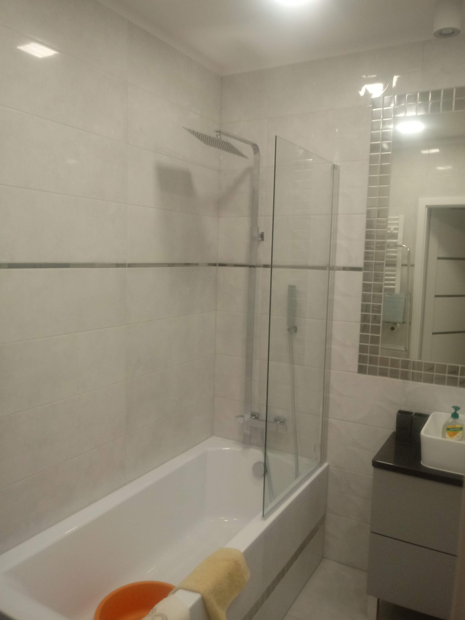 Biała łazienka z wanną, szklanym parawanem nawannowym i deszczownicą, wykończona płytkami w jasnych odcieniach szarości z dekoracyjnym pasem mozaiki. W lustrze odbija się fragment grzejnika...