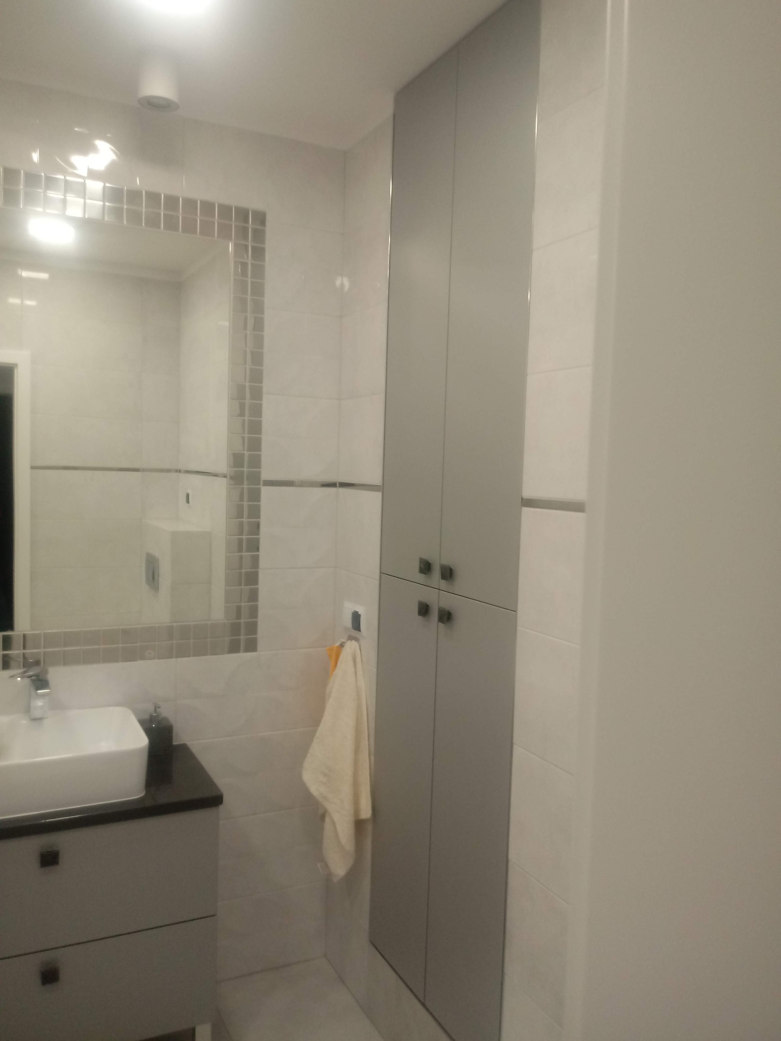 Nowoczesna łazienka w odcieniach szarości i bieli z dużym lustrem obramowanym mozaiką, wiszącą szafką z umywalką i wysoką szafą, uzupełniona beżowym ręcznikiem.