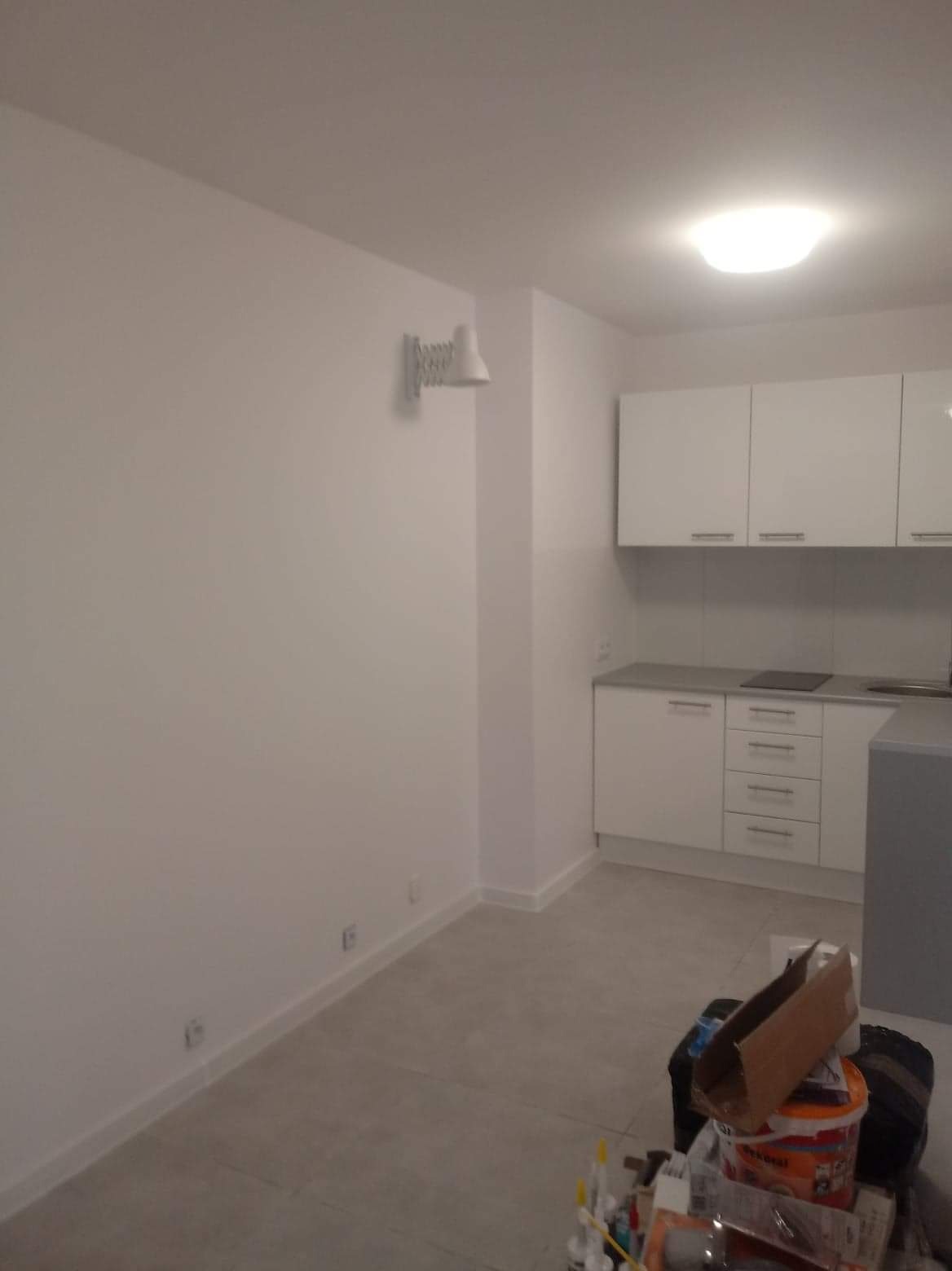 Pomalowane na biało wnętrze mieszkania z widoczną ścianą, sufitem, podłogą z szarymi płytkami, lampą na ścianie i fragmentem zabudowy kuchennej z białymi szafkami; na podłodze widoczne materiały...