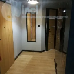 Wykończony korytarz z drewnianą podłogą, drzwiami o fornirze drewnopodobnym, lustrem w czarnej ramie i nowoczesną lampą sufitową; widoczny fragment ściany z okładziną imitującą marmur.