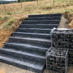 Schody zewnętrzne z ciemnej kostki kompozytowej, zabezpieczone gabionami z kamienia łamanego, widok z dołu na tle trawiastych zboczy.