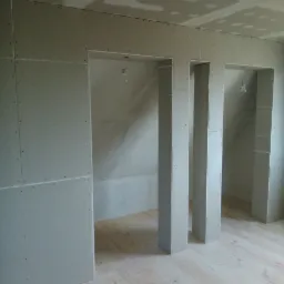 Wnętrze w trakcie montażu płyt kartonowo-gipsowych na ścianach i suficie, widoczne trzy otwory drzwiowe z obudowanymi słupkami, surowa drewniana podłoga.