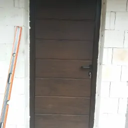Brązowe, segmentowe drzwi garażowe z kluczem w zamku, zamontowane w surowym murze z białych bloczków, poziomica oparta o ścianę.