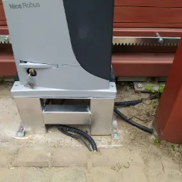 Szary automat do bramy przesuwnej Nice Robus zamontowany na metalowej podstawie na kostce brukowej, widoczne podłączenia kablowe i zębatka bramy.