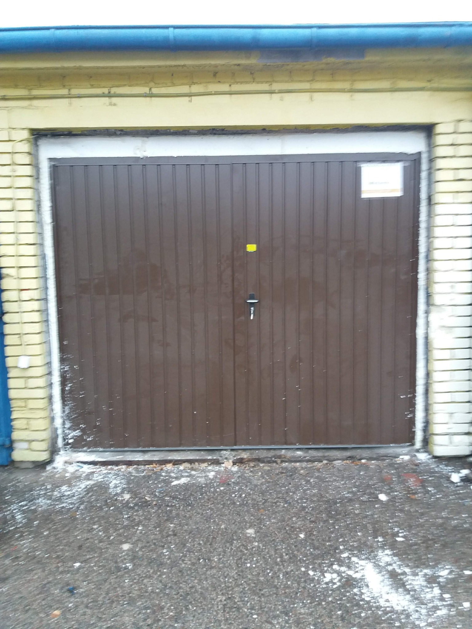 Zewnętrzny widok brązowej, segmentowej bramy garażowej z widocznymi śladami uszczelniania i niewielkimi zabrudzeniami w dolnej części, osadzonej w żółtej ceglanej ramie.