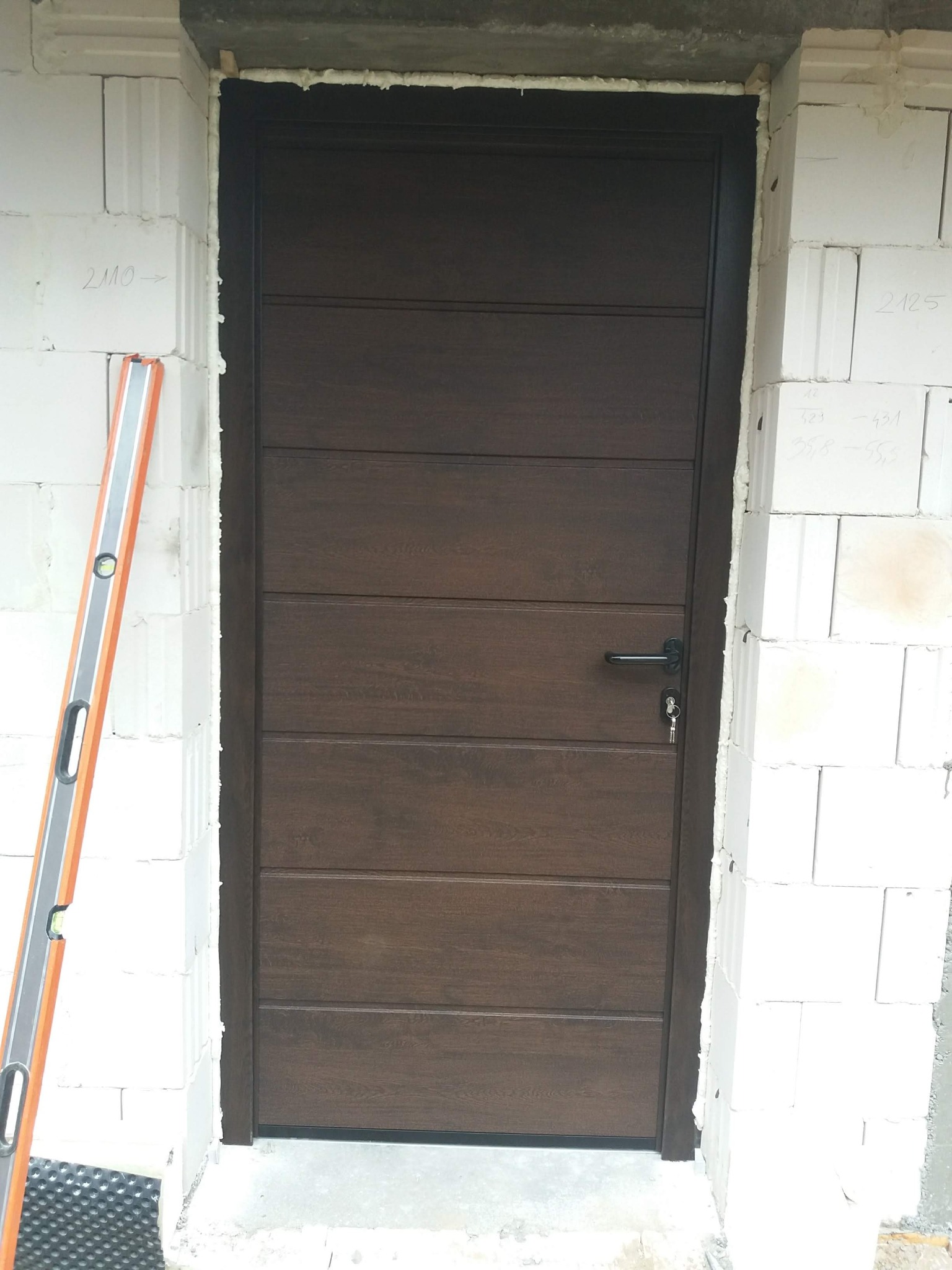 Brązowe, segmentowe drzwi garażowe z kluczem w zamku, zamontowane w surowym murze z białych bloczków, poziomica oparta o ścianę.