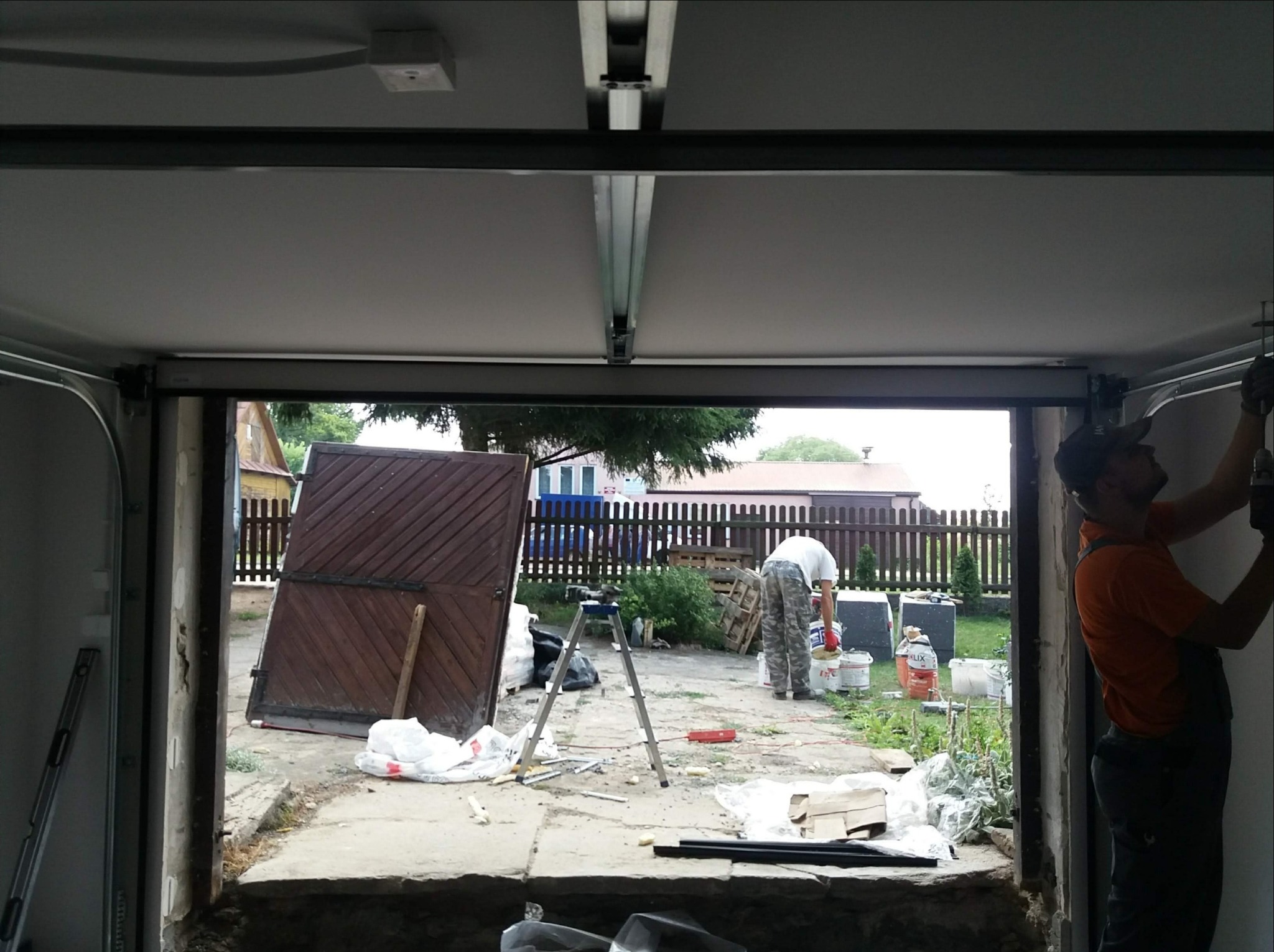 Montaż bramy garażowej w Krupnikach: widok z wnętrza garażu na podwórko, gdzie widać narzędzia, drabinę i drugiego pracownika przygotowującego materiały. Jeden z monterów w pomarańczowej koszulce...