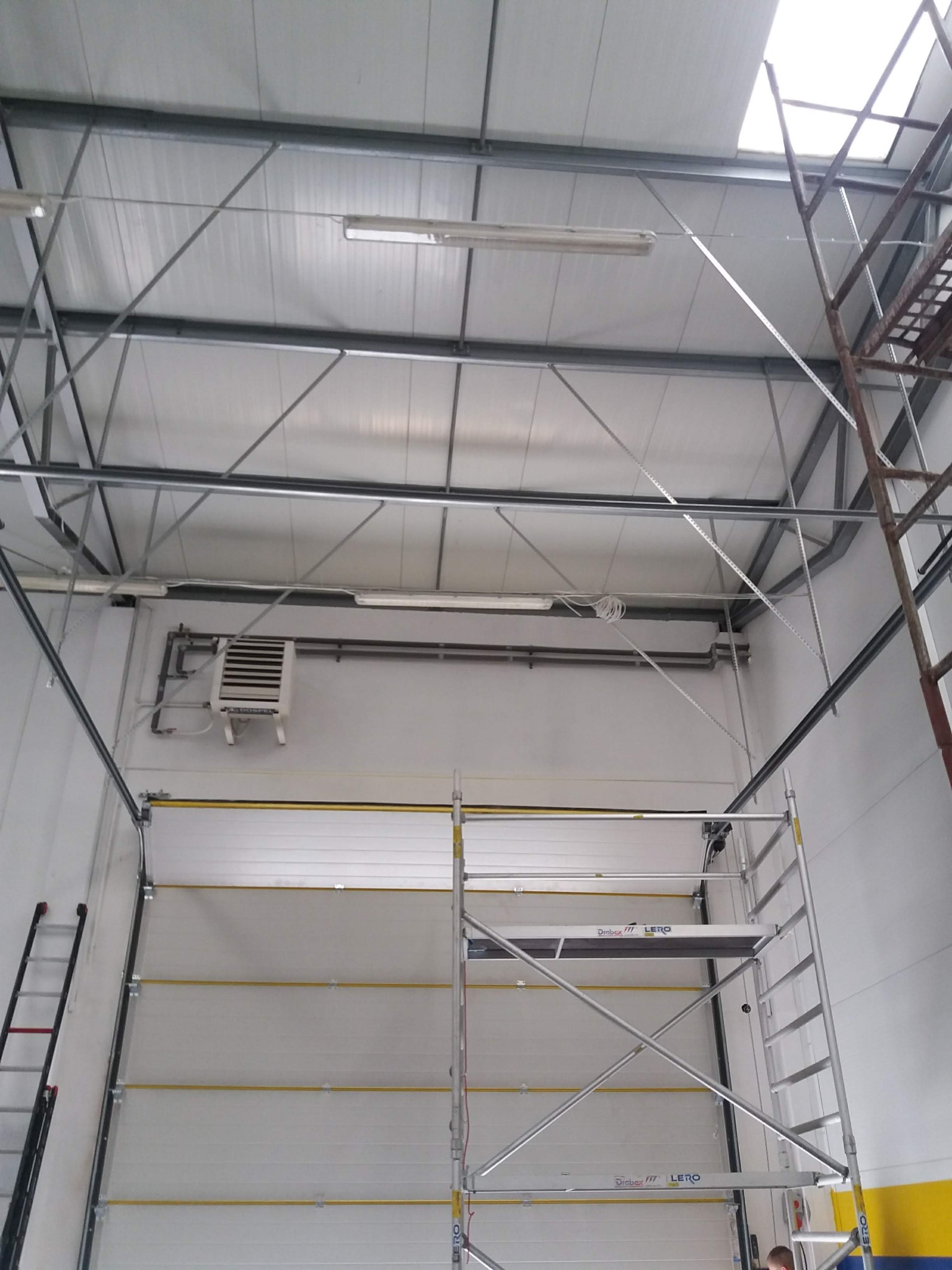 Wnętrze hali przemysłowej z widoczną bramą segmentową, grzejnikiem przemysłowym i rozstawionym rusztowaniem aluminiowym.
