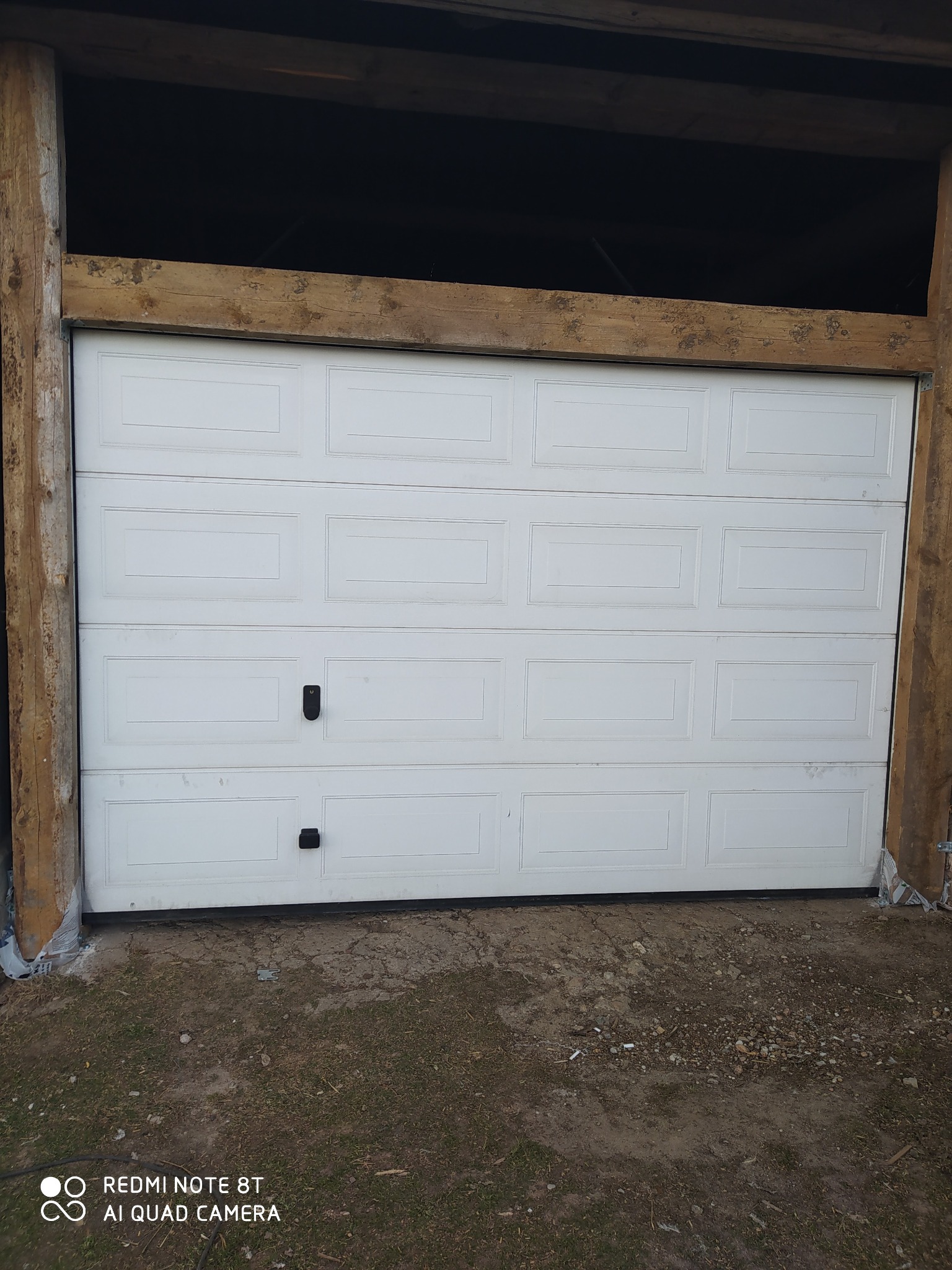 Zamknięta biała segmentowa brama garażowa z czarnymi uchwytami, osadzona w drewnianej konstrukcji z surowego drewna. Podłoże ziemne.