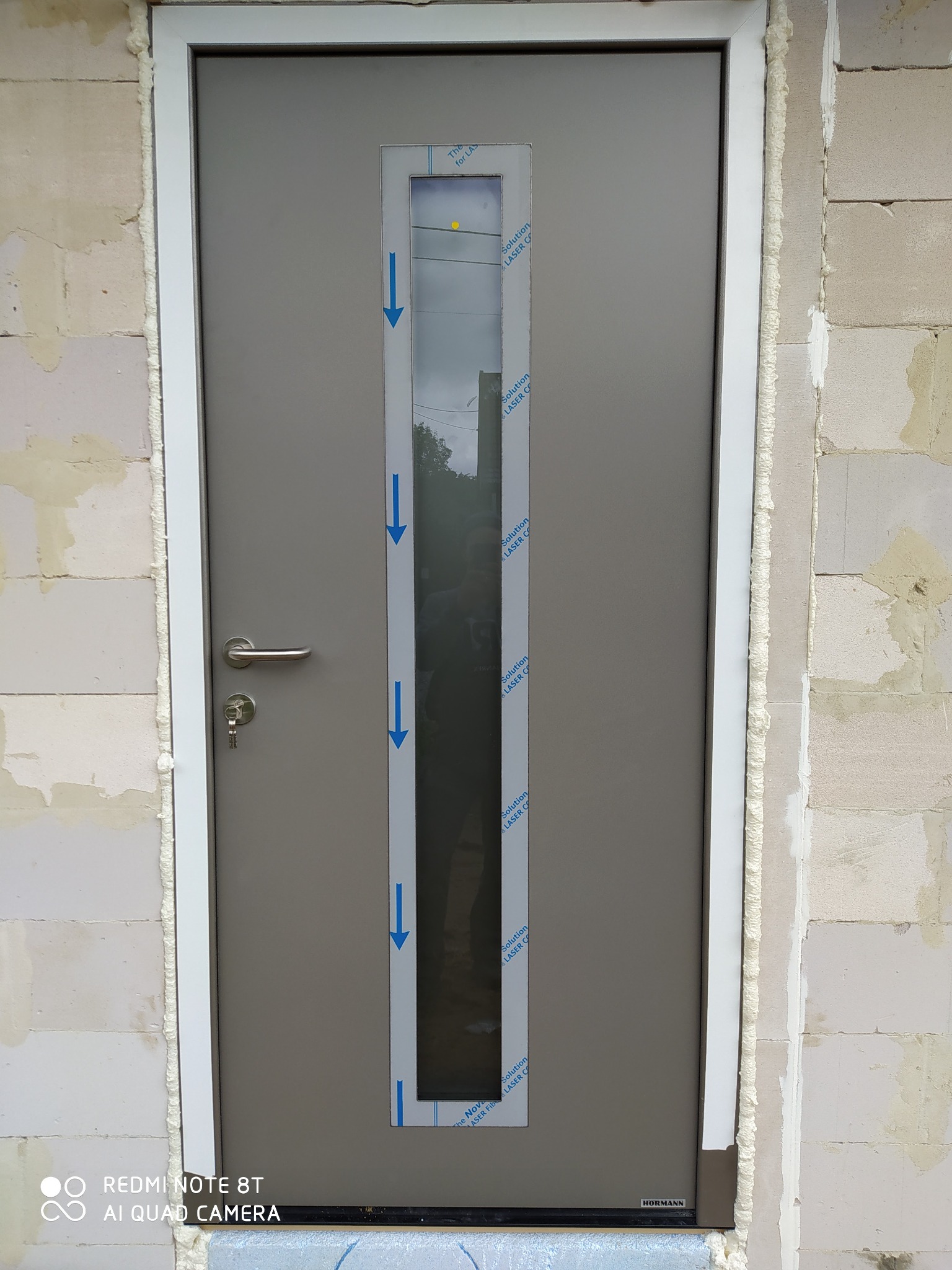 Nowo zamontowane szare drzwi wejściowe z długim, wąskim oknem, zabezpieczone folią ochronną z niebieskimi strzałkami, osadzone w surowej ścianie z pustaków, uszczelnione pianką montażową.