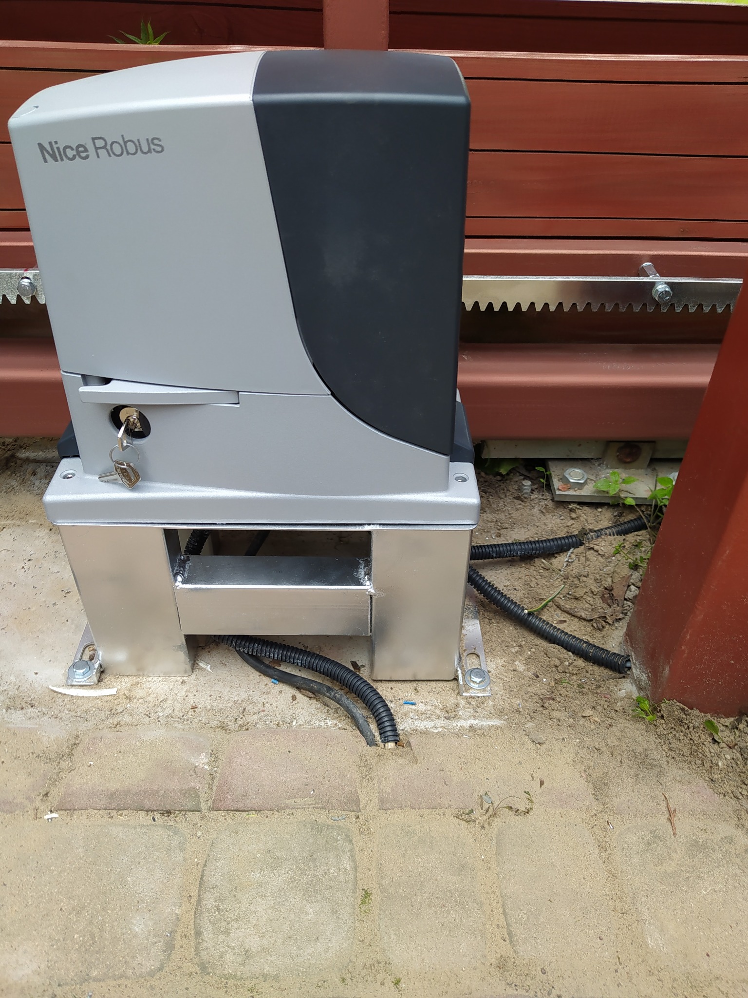 Szary automat do bramy przesuwnej Nice Robus zamontowany na metalowej podstawie na kostce brukowej, widoczne podłączenia kablowe i zębatka bramy.