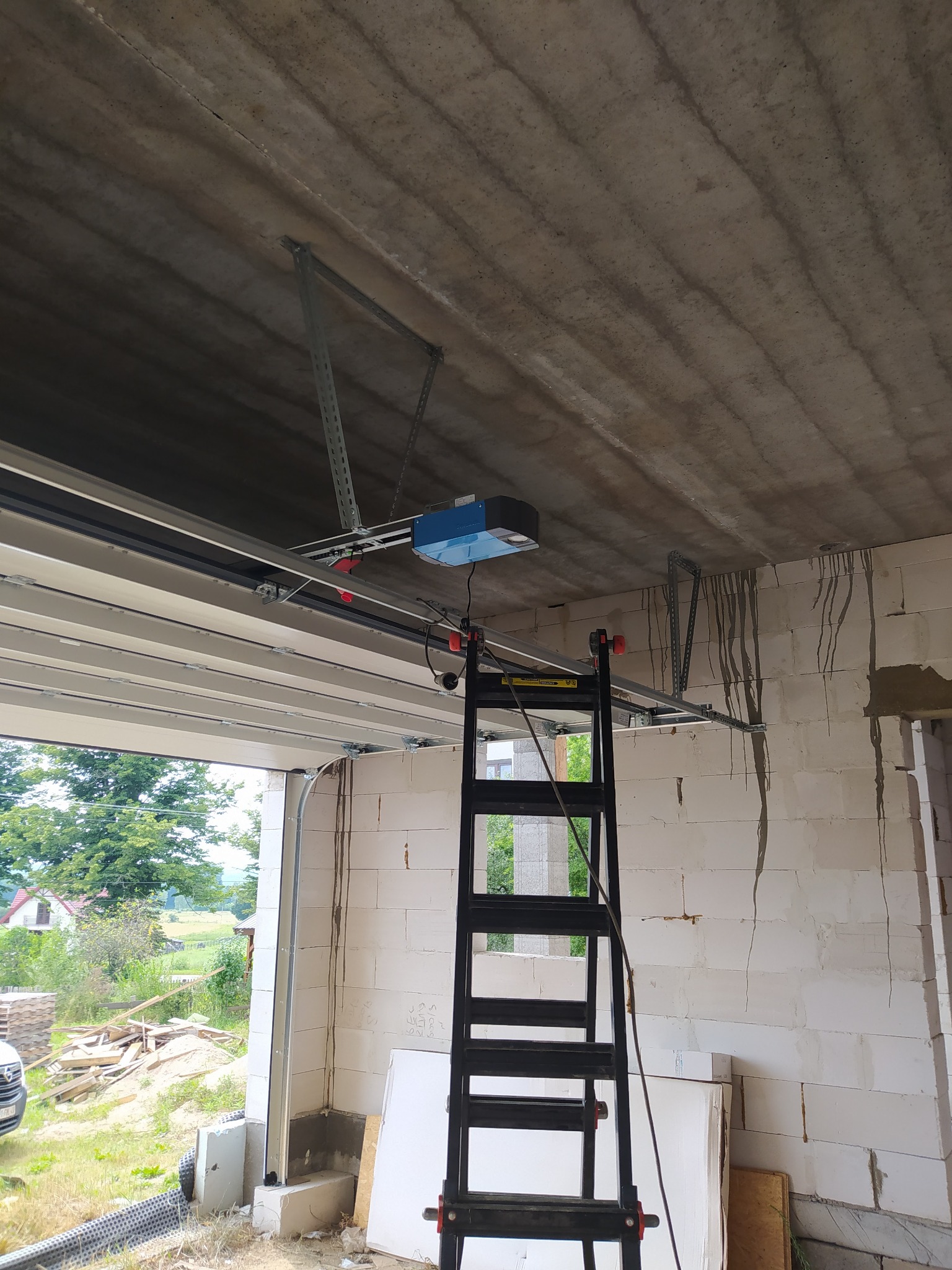 Montaż automatyki do bramy garażowej segmentowej, widoczne szyny, silnik i drabina w surowym wnętrzu garażu z widokiem na zewnątrz.