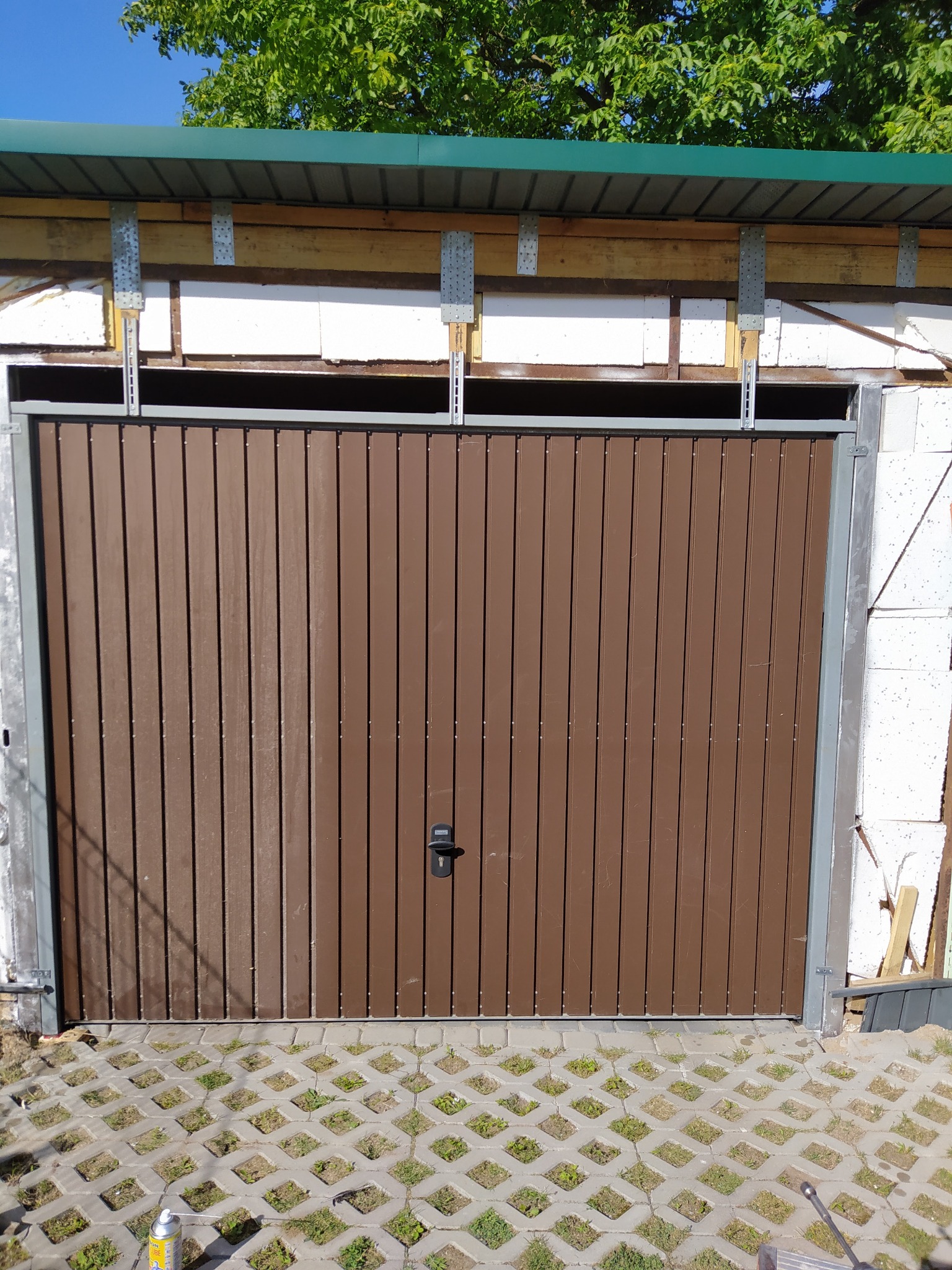 Brązowa brama garażowa z pionowymi panelami, widoczne mocowania do ściany z pustaków, podłoże z ażurowych płyt betonowych, puszka z pianką montażową w rogu.