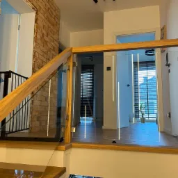 Nowoczesne schody z drewnianymi stopniami i szklaną balustradą, oświetlone pionowymi paskami LED, widok na jasne wnętrze z roletami.