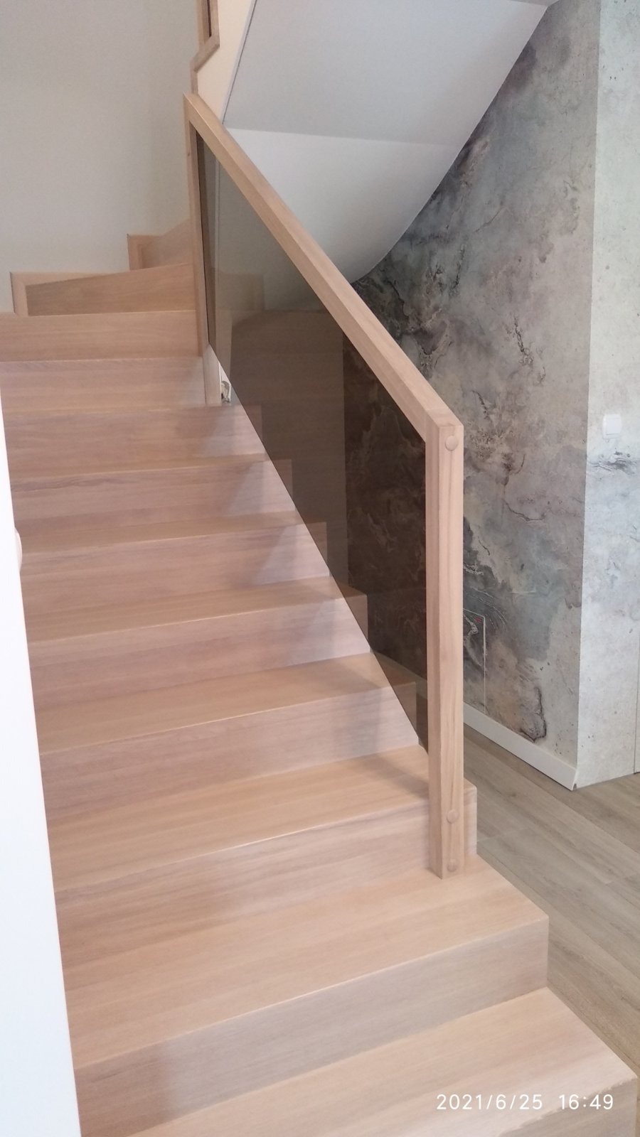 Jasne, proste schody z drewnianymi stopniami i szklaną balustradą, widok z dołu, marmurkowa ściana w tle.