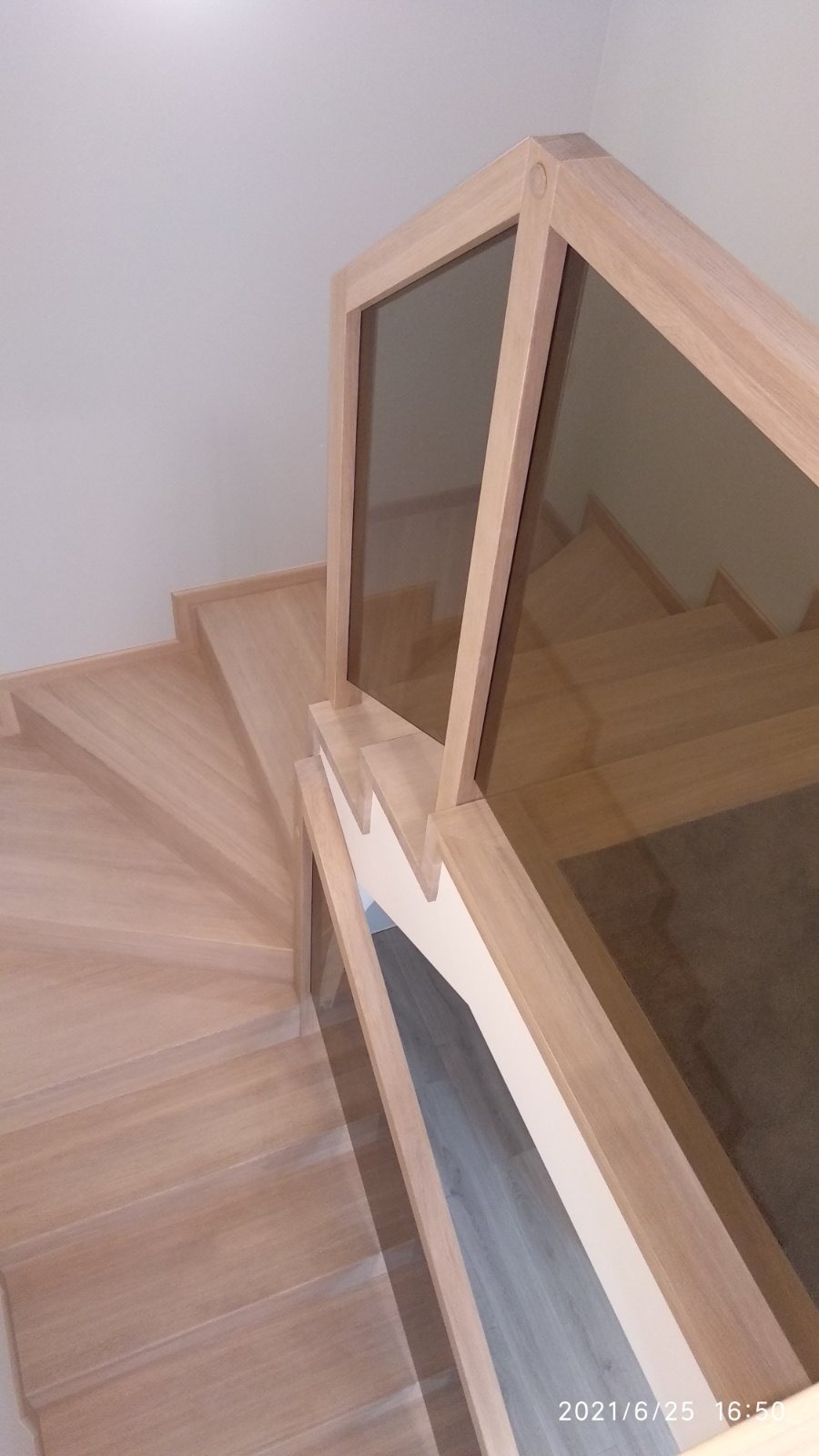 Widok z góry na jasne, spiralne schody z drewna z balustradą ze szkła w drewnianej ramie, widoczny fragment podłogi pod schodami.