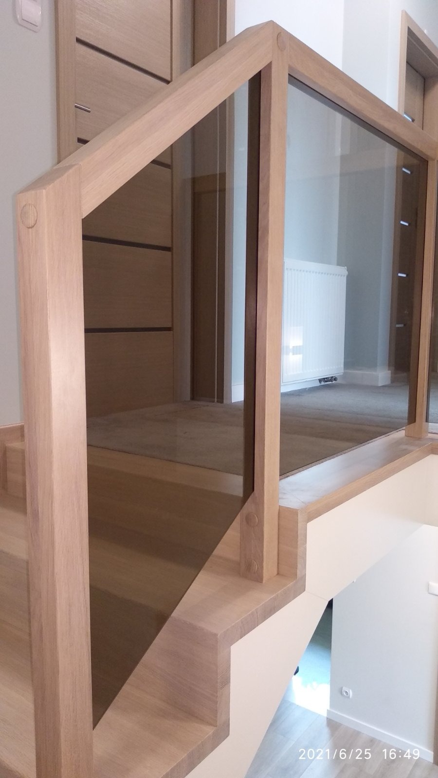 Fragment nowoczesnych schodów z jasnego drewna z przeszkloną balustradą, widok z dołu na pierwsze stopnie i fragment korytarza w tle.