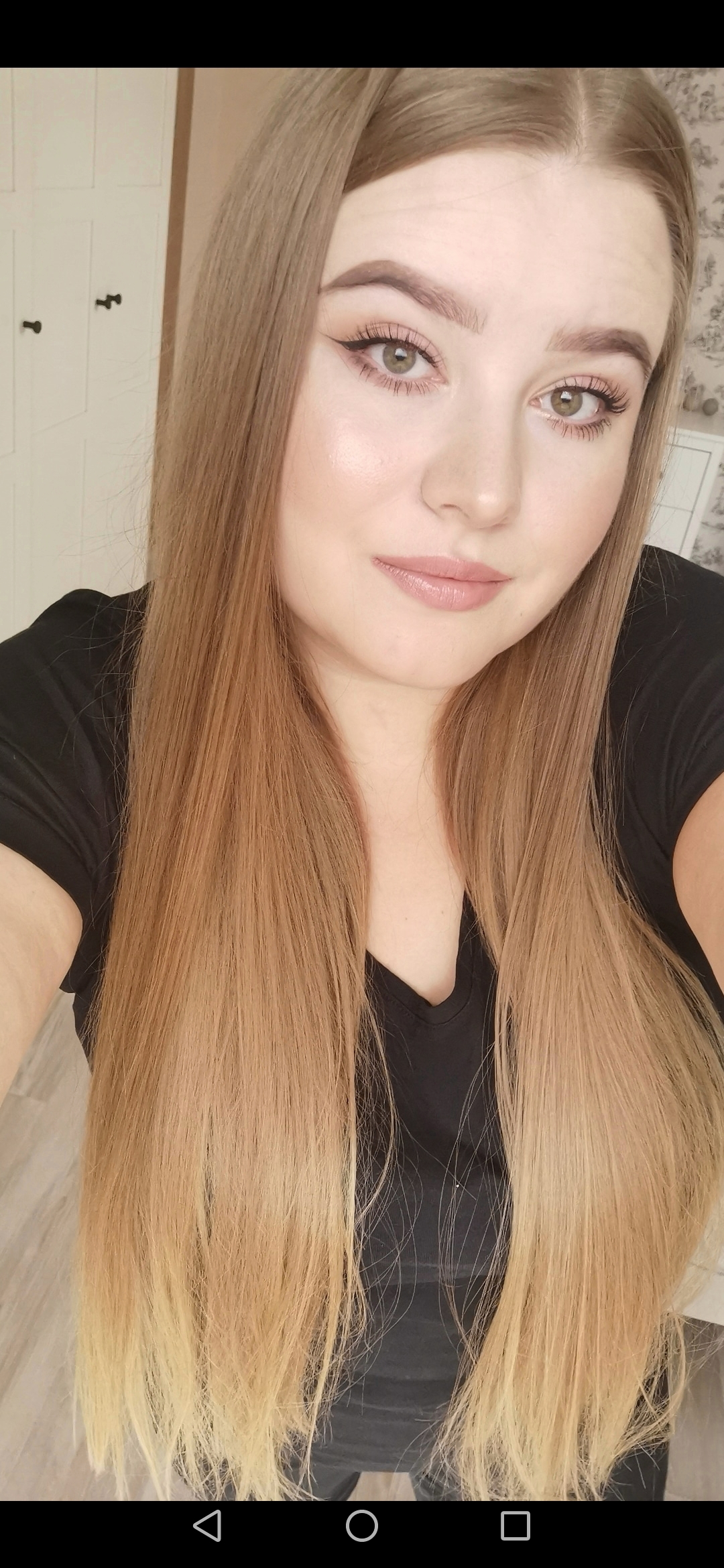 Portret młodej kobiety z długimi, blond włosami z ombre, delikatny makijaż z czarną kreską na powiece, na tle białych drzwi.