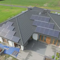Widok z góry na dom z szarą dachówką, na której zamontowano panele fotowoltaiczne; widoczne są również elementy ogrodu i otaczające pola.